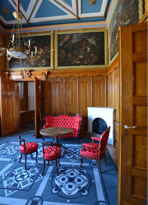 Blick in den Kaminsaal mit holzgetäfelten Wänden, großformatigen Gemälden und einer roten gepolsterten Sitzgruppe im Rokoko-Stil auf blau gemustertem Ornament-Teppich.