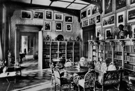 Historic photograph of a salon with half-height bookshelves and paintings and sculptures above, and ornate upholstered chairs around a round table. The parquet floor is laid in a chequerboard pattern.