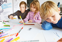 Three children are sitting at a table and drawing. Colourful drawings are spread out on the table, with colourful felt-tip pens on top.