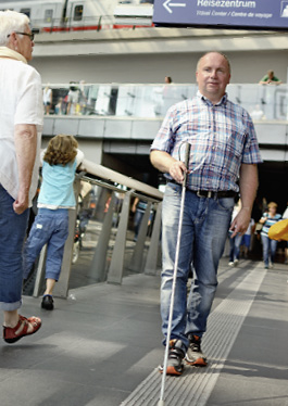 Foto eines blinden Mannes, der sich in einem Bahnhof mit Blindenlangstock am Bodenleitsystem orientiert