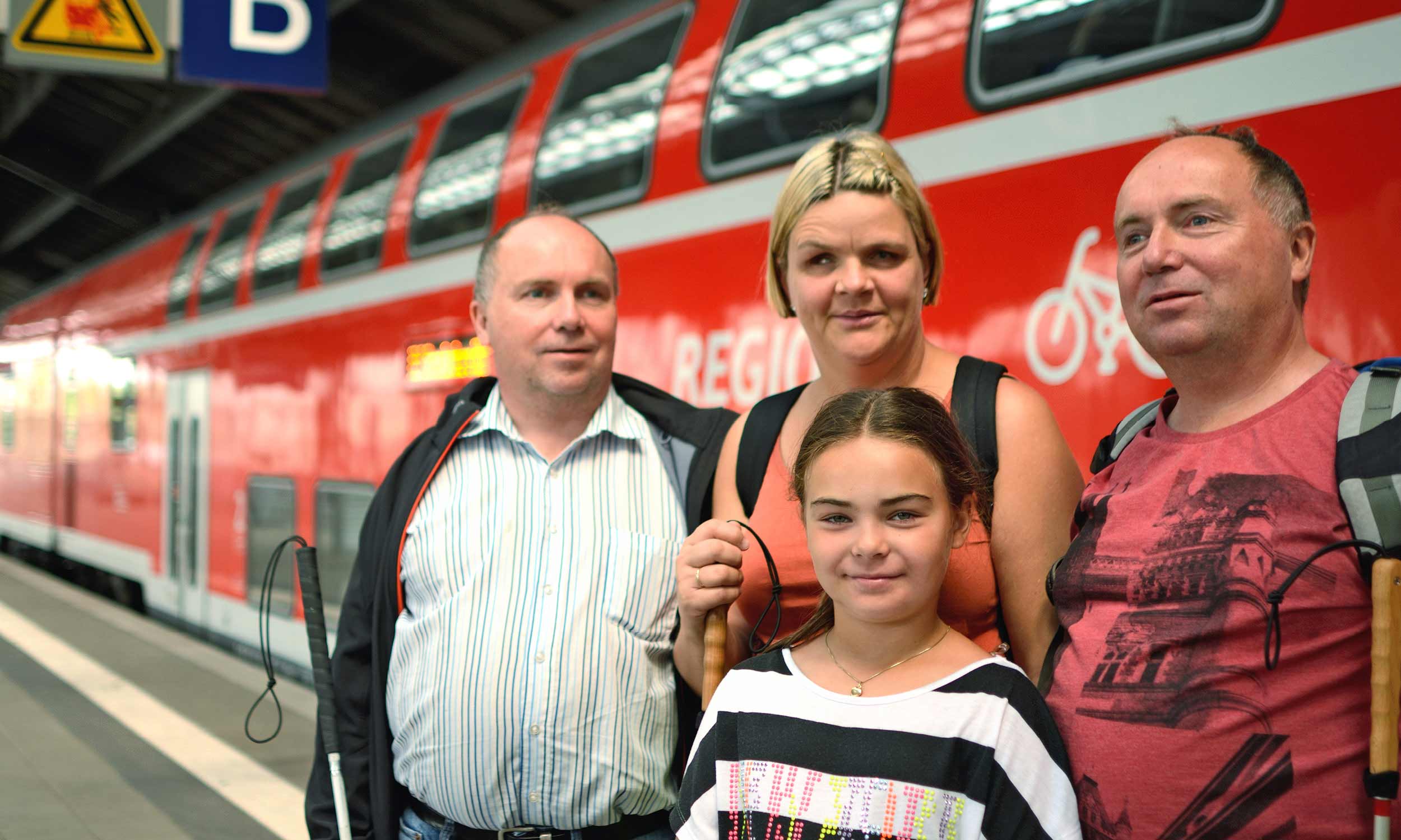 Group photo of tourists in Berlin - two adult blind brothers and a blind wife with a sighted daughter. They are standing on the platform in front of a red double-decker regional express.