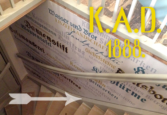 The staircase in the exhibition Klosteramt Dobbertin looking down from above. The staircase hatch is framed with a white grille. The wooden steps lead along a wall decorated with yellow, blue and grey historical lettering. A curved white banister is mounted on the wall.