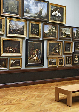 Photo showing a view into an exhibition hall of the Staatliches Museum Schwerin. On a light grey wall hang many paintings of Dutch painting of the 17th century in massive golden and dark frames. At some distance from the wall is a wooden bench on the parquet floor.