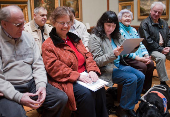 Photo of a test group of blind and visually impaired people in an exhibition hall of the State Museum Schwerin. Three women are sitting in the front row, framed by two men. In the row of seats behind them are two more people. They are checking Braille prints, relief transparencies and relief prints of our painting guide for legibility. Obviously with success, as can be seen from their faces.