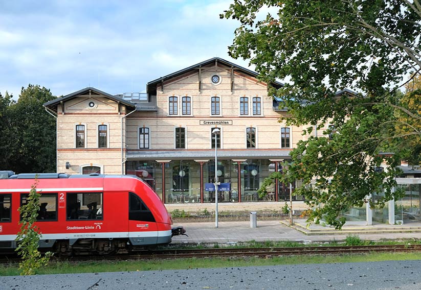 Außenansicht des Bahnhofs Grevesmühlen. Der Bahnsteig mit einem roten Zug im Vordergrund. Im Hintergrund sehen wir den Bahnhofs mit der Glasfassade des Wintergartens.