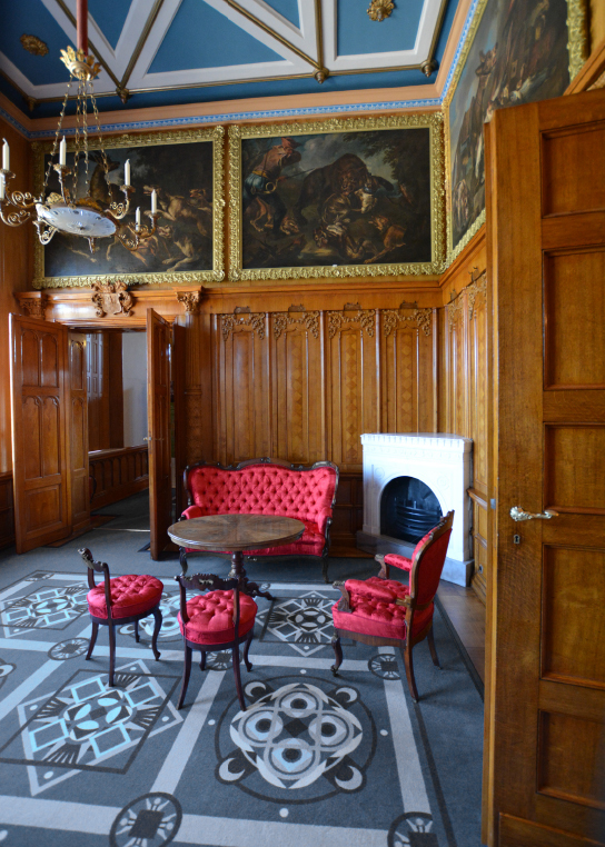 Blick in den Kaminsaal mit holzgetäfelten Wänden, großformatigen Gemälden und einer roten gepolsterten Sitzgruppe im Rokoko-Stil auf blau gemustertem Ornament-Teppich.
