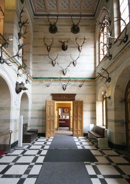 Photo of the entrance and trophy hall. The walls of the two-storey high hall are adorned with antlers of various game species. The mammoth floor, structured in grey and white rectangles, is covered by a plain grey runner.