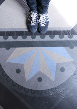 Detail of a carpet design. The pattern shows a two-coloured star in light blue and grey in a dark grey circle framed by blue-grey dots. Further lines and dots extend the pattern. The feet of a person in blue-grey trainers can be seen on the upper edge of the picture.