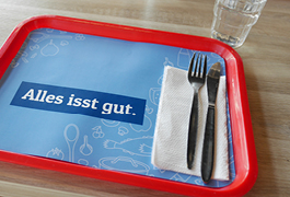 Foto eines Speisetabletts mit Besteck auf hellblauer Papierunterlage mit dem Slogan „Alles isst gut“.
