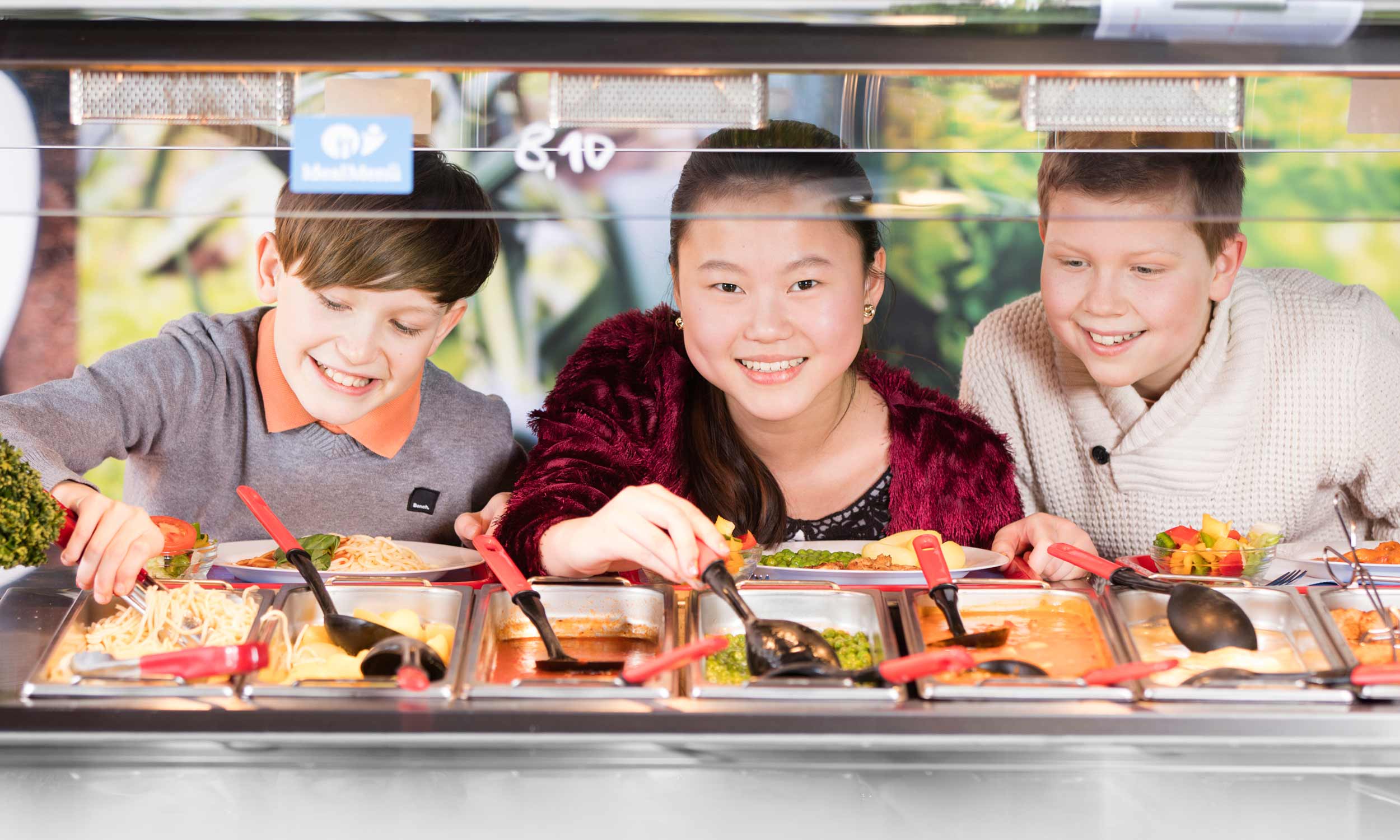 Foto mit drei Schülern, die freudig an der Selbstbedienungstheke ihr Mittagessen zusammenstellen