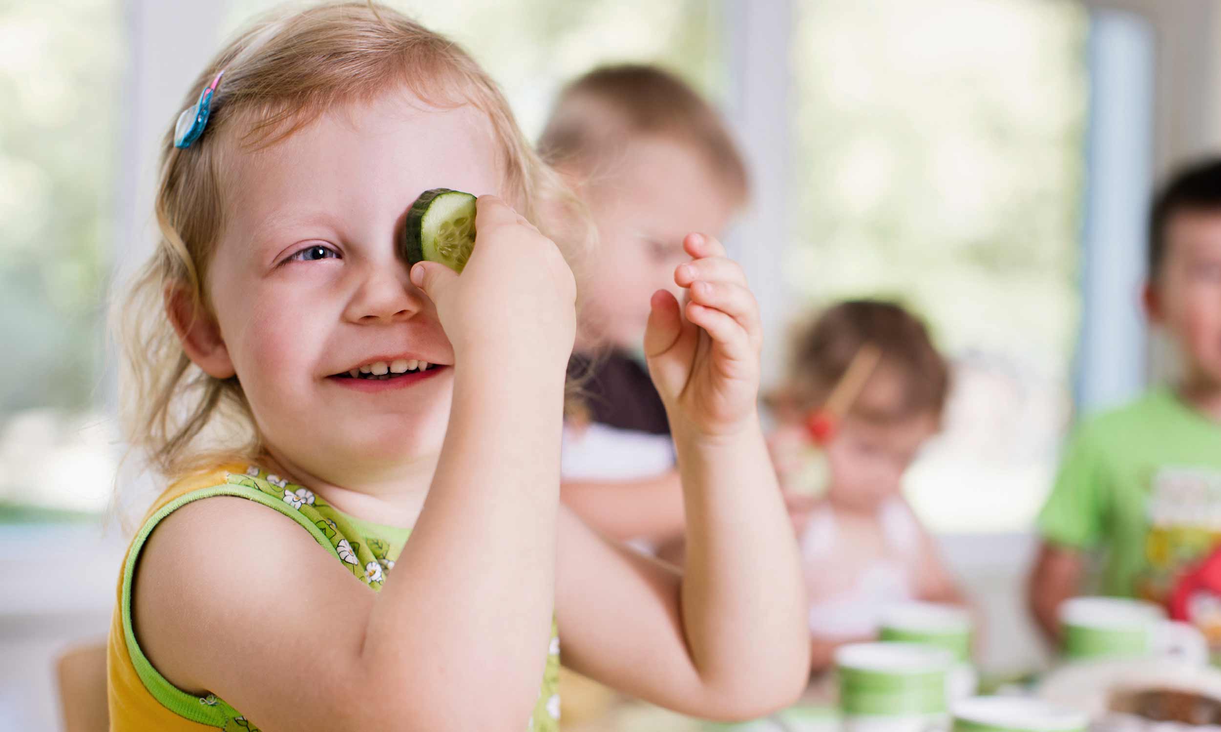 Photo of a happy nursery child at the table covering one eye with a slice of cucumber