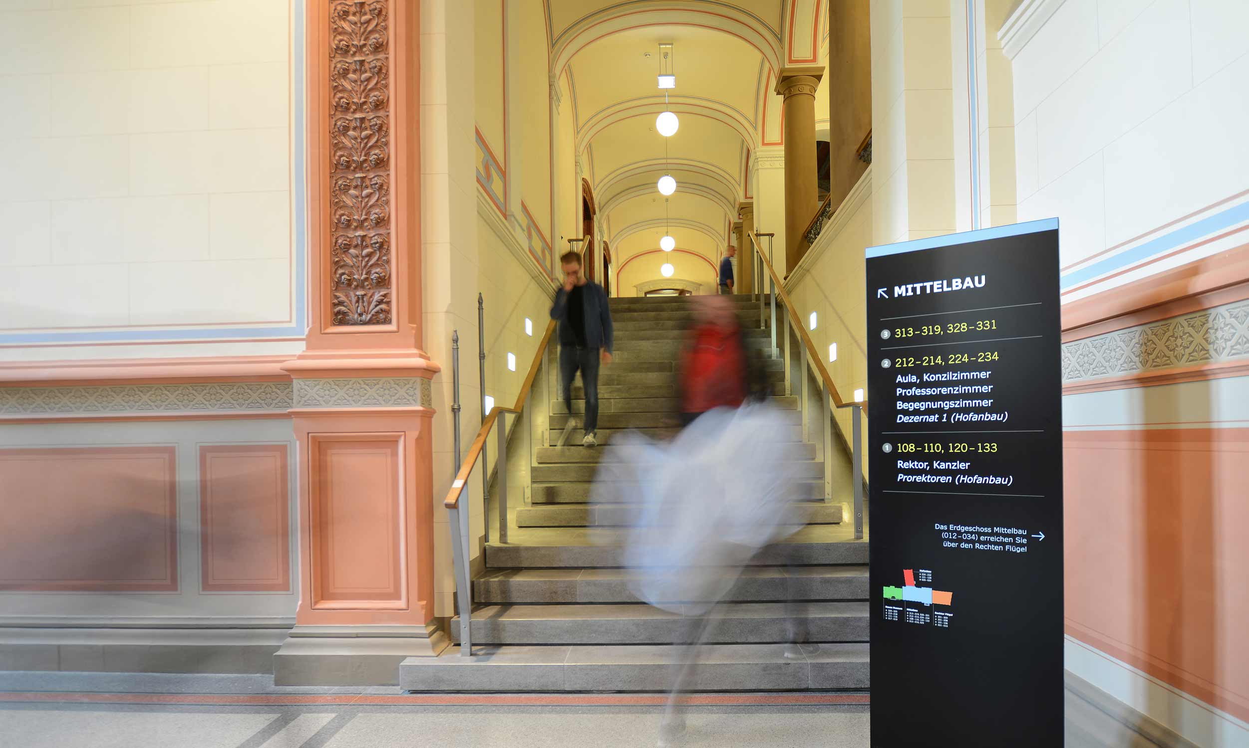 A view into the entrance area of the University of Rostock. In the background of the picture, the staircase leading to the first floor, with a few people in motion out of focus. On the right you can see a black stand with light-coloured lettering, an element of the signposting system for the University of Rostock.