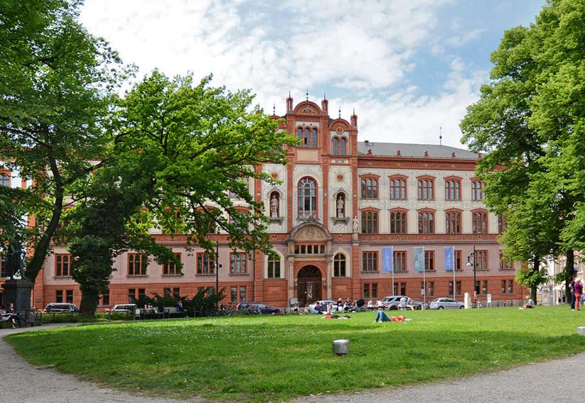 Eine Außenansicht des Hauptgebäudes der Universität Rostock mit Blick auf den Haupteingang. Es ist ein prachtvolles Gebäude im Stil der mecklenburgischen Neorenaissance. Im Vordergrund befindet sich eine Wiese. Bäume mit grüner Krone verdecken leicht die Sicht auf das Gebäude.