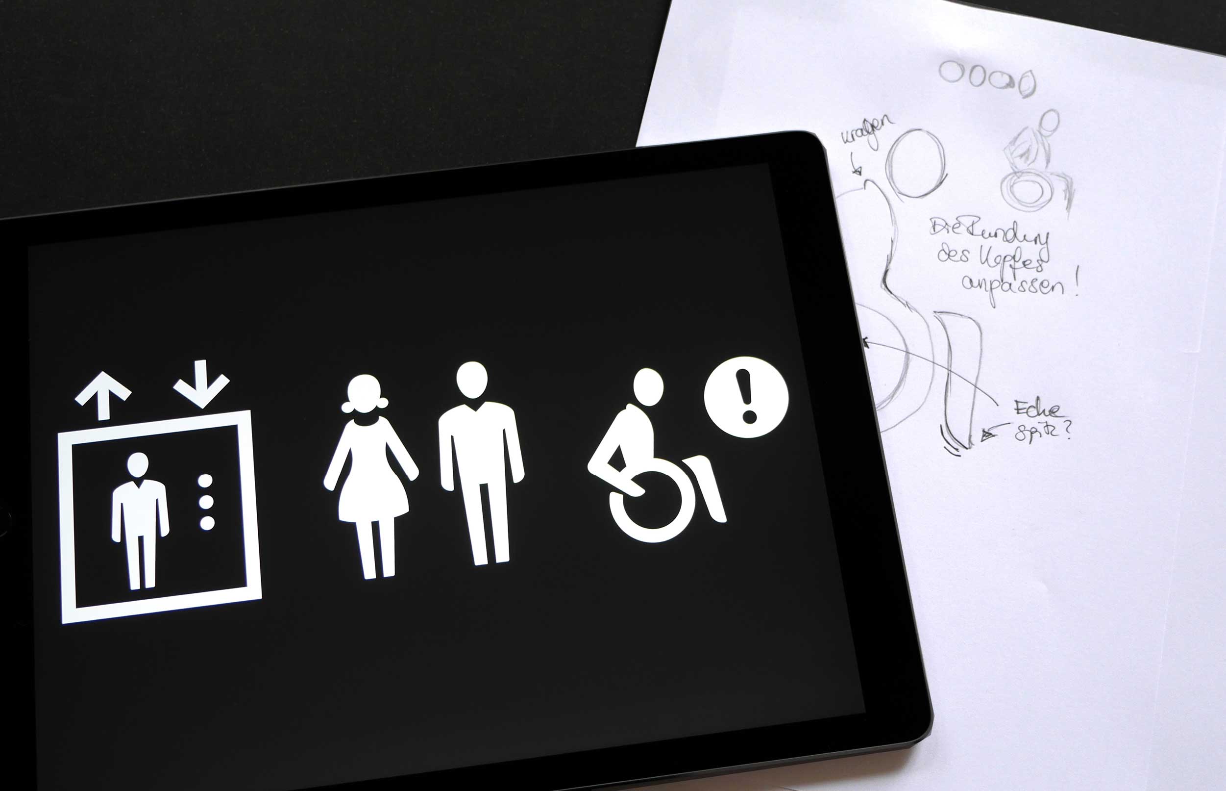 Auf einem iPad werden verschiedene weiße Piktogramme auf schwarzem Untergrund gezeigt. Es handelt sich um die Piktogramme zur Kennzeichnung des Fahrstuhls, der Damen- und Herren-Toiletten sowie der barrierefreien Toiletten, die mit einem Rollstuhlfahrer kenntlich gemacht werden. Vom I-Pad leicht verdeckt werden Skizzen zum Rollstuhlfahrer-Piktogramm.