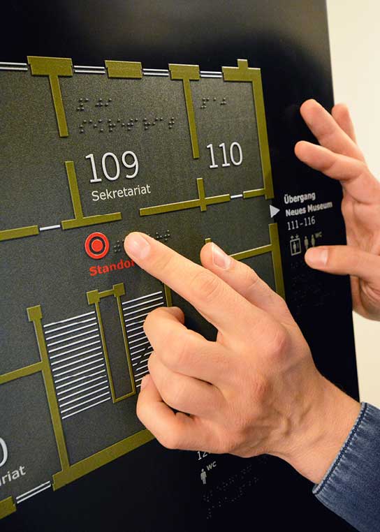 A tactile floor overview in vertical position. A person uses a finger to feel the braille lettering indicating the location.