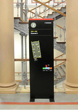 Photo of a display stand on the second floor. The display includes a tactile floor overview and a graphic of the individual building sections. It stands in the middle of two cream-colored stone columns. The building's stairwell is visible in the background.