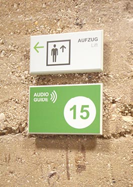Ein Hinweisschild zum Aufzug in weiß mit schwarzem Piktogramm. Darunter ein grünes Schild mit der Nummer für den Audio Guide