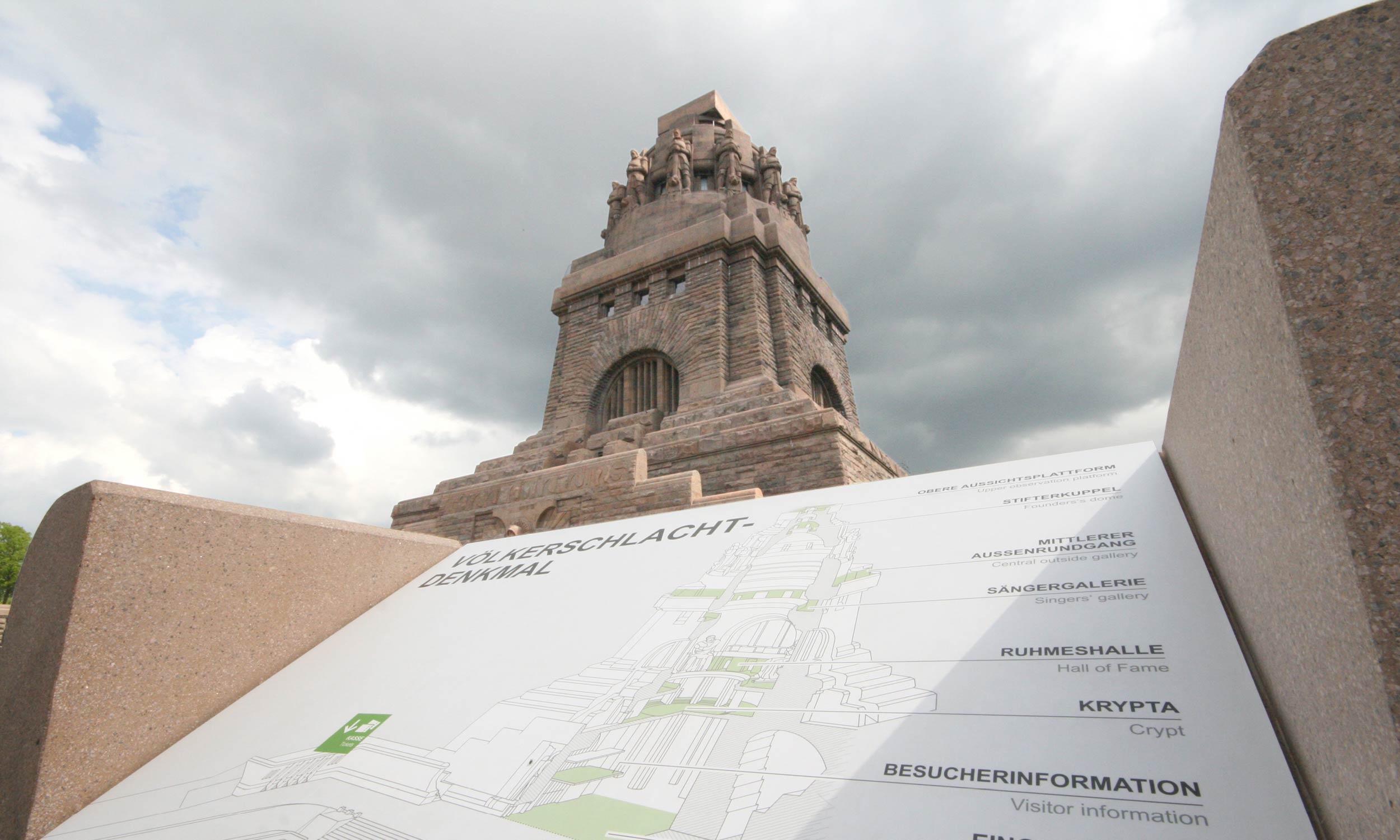 Fokus auf eine Infotafel, die das Denkmal im Querschnitt zeigt. Im Hintergrund ist das imposante Denkmal zusehen, das sich in den bewölkten Himmel streckt