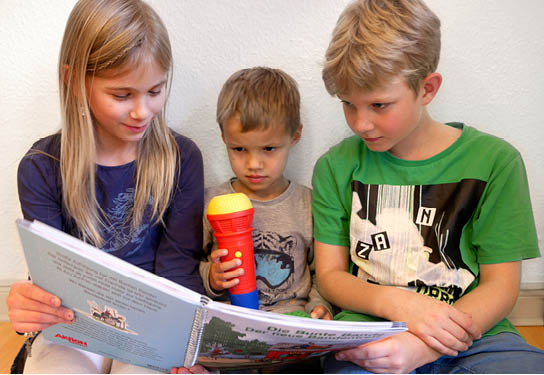 A girl and two boys are sitting side by side on the floor with their backs against a wall. The girl reads the stories of the colourful gang to the two boys.