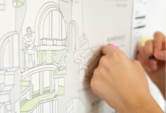 A picture from the design process. A detailed drawing of the monument in cross-section hangs on the wall. Someone is describing notes stuck to the drawing.