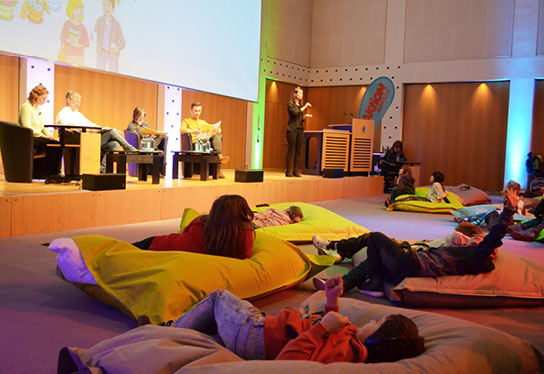 A cosy listening room can be seen. The book "Die Bunte Bande" (The Colourful Gang) is being read aloud on the stage. On the floor in front of it, children sit and lie on colourful cushions and listen intently.