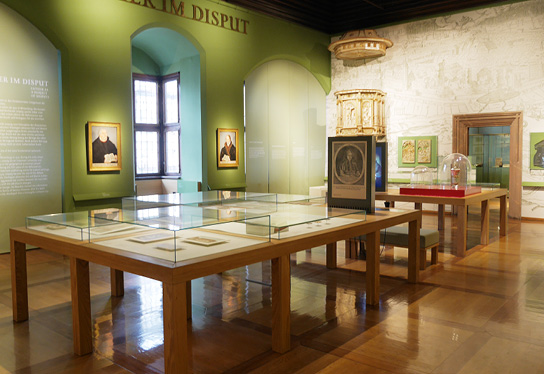 Foto des Ausstellungsraumes „Luther im Disput“ im Stadtgeschichtlichen Museum Leipzig mit diversen Vitrinen und Exponaten