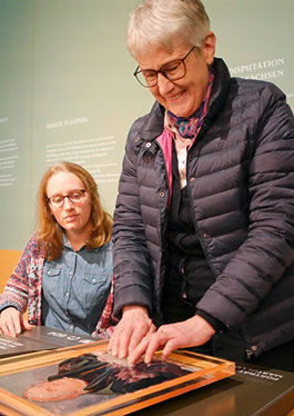 Foto einer stehenden blinden Frau, die das in die Taststation integrierte Luther-Gemälde ertastet