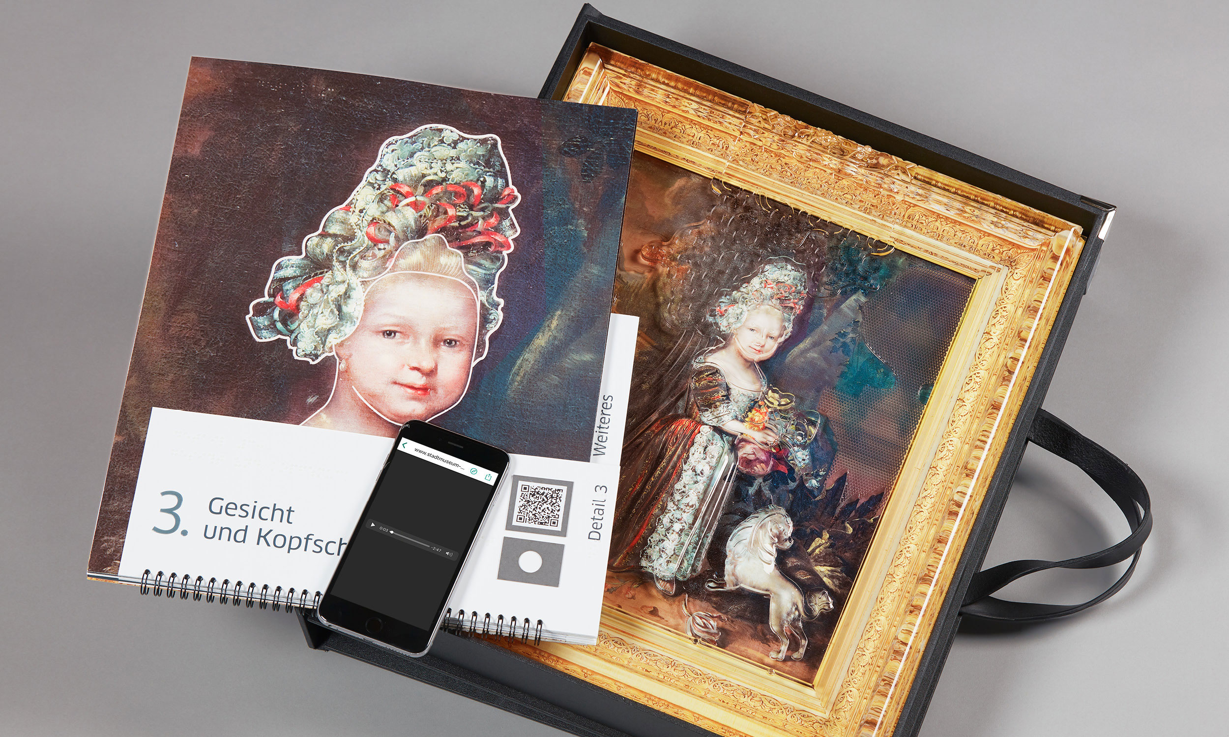 The photo shows the tactile painting of the "Little Princess" in a custom-made case, the opened lid of which serves as a slanted desk. On the left side of the tactile painting is the open booklet accompanying the painting, which covers part of the tactile painting. A detailed illustration is open, as well as the page with the matching inscription in Braille and black lettering, and the QR code and the label for the hearing pen are also visible. On top of the accompanying booklet is a smartphone whose screen shows that an audio file is being played.