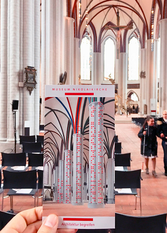 Dieses Foto zeigt den Flyer für die Dauerausstellung „Architektur begreifen“; im Hintergrund befindet sich der Innenraum der Kirche.