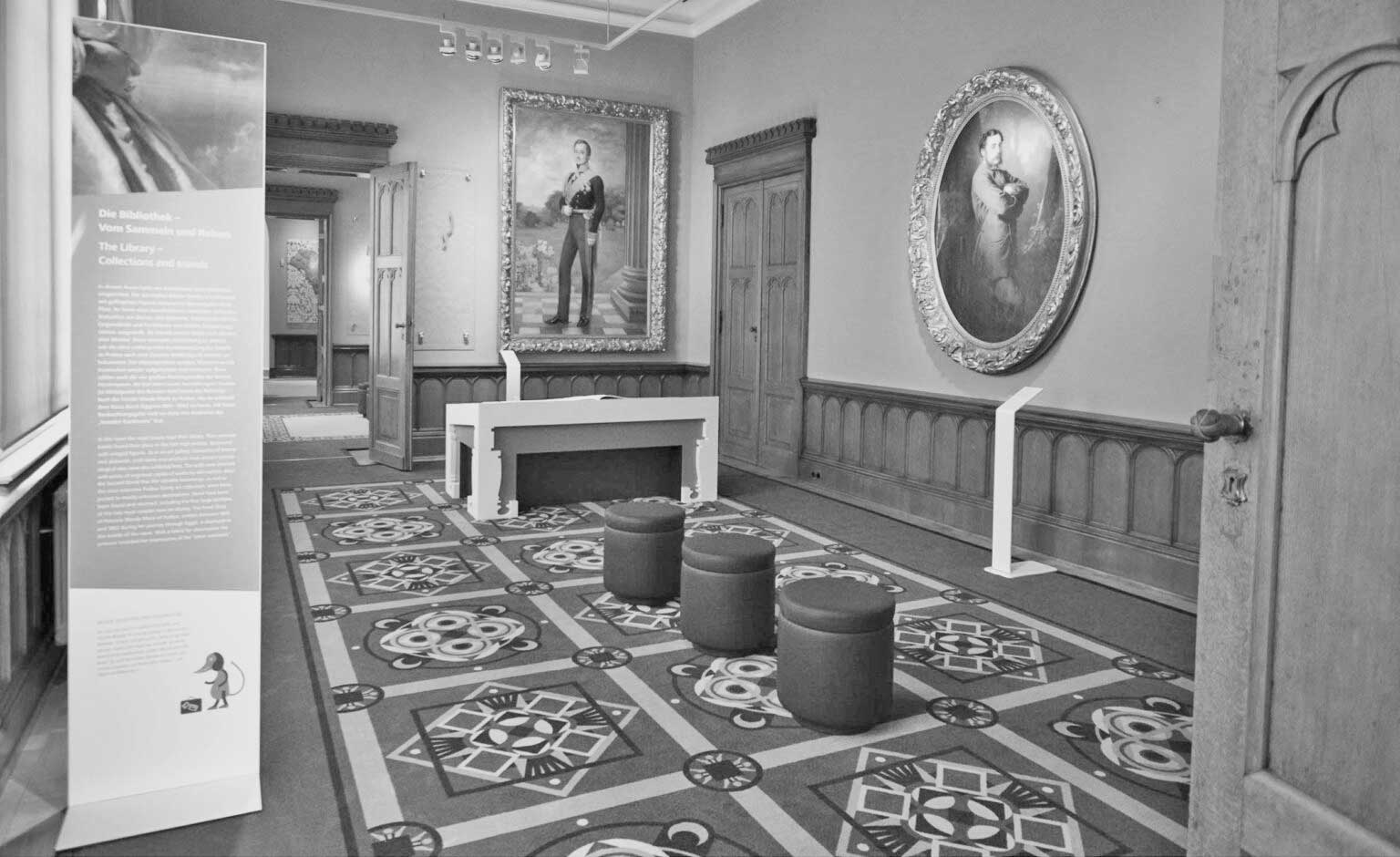 Photo of the next project: Granitz hunting lodge: It presents the contemporary image of a salon with a wood-panelled ceiling and honey-coloured double doors. The eye-catcher is the ornamental carpet in shades of grey that covers the floor of the room. In the centre of the picture, three dark red stools are placed in a row. On the wall in the background of the picture hangs a large rectangular painting of a nobleman in a gold frame, with a modern white table below. On the wall to the right of the picture hangs a portrait of a lady in a pompous oval gold frame.