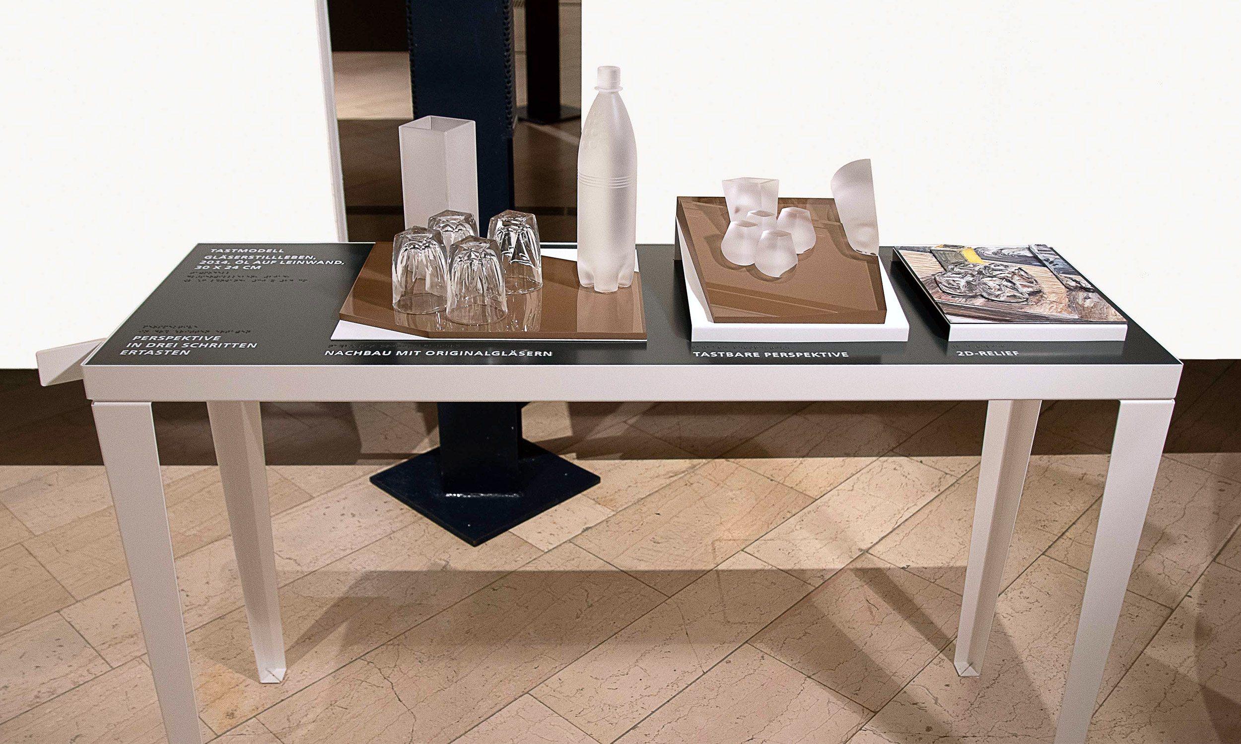 Photo of the three tactile models of the painting "Glass Still Life" on a table. On the far left info text, then the three-dimensional model of the painting, next to it a perspective-distorted three-dimensional view and on the far right a two-dimensional image of the painting with tactile lines.