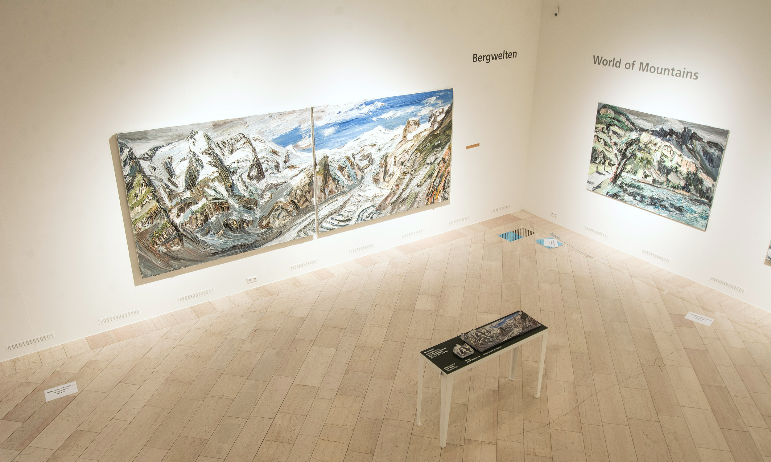 View from above into a corner of the exhibition hall. On the left wall the very large, two-part painting "Bergwelten". In front of it on a table the tactile model to the painting.
