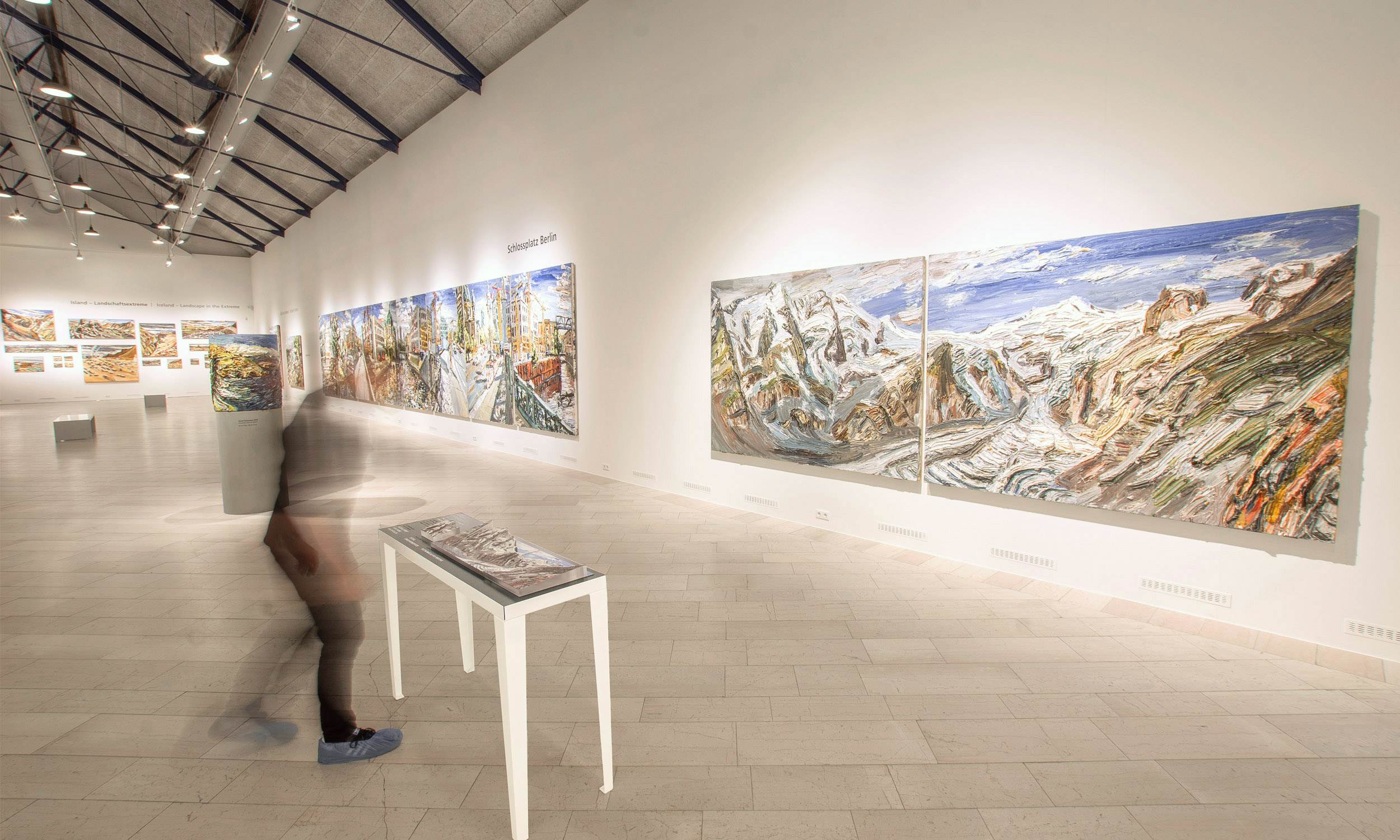 View into a long exhibition hall. On the right on the walls the paintings of the artist. At the very front the painting "Bergwelten", in front of which the corresponding tactile model can be seen on a table. At the table, out of focus, a person.