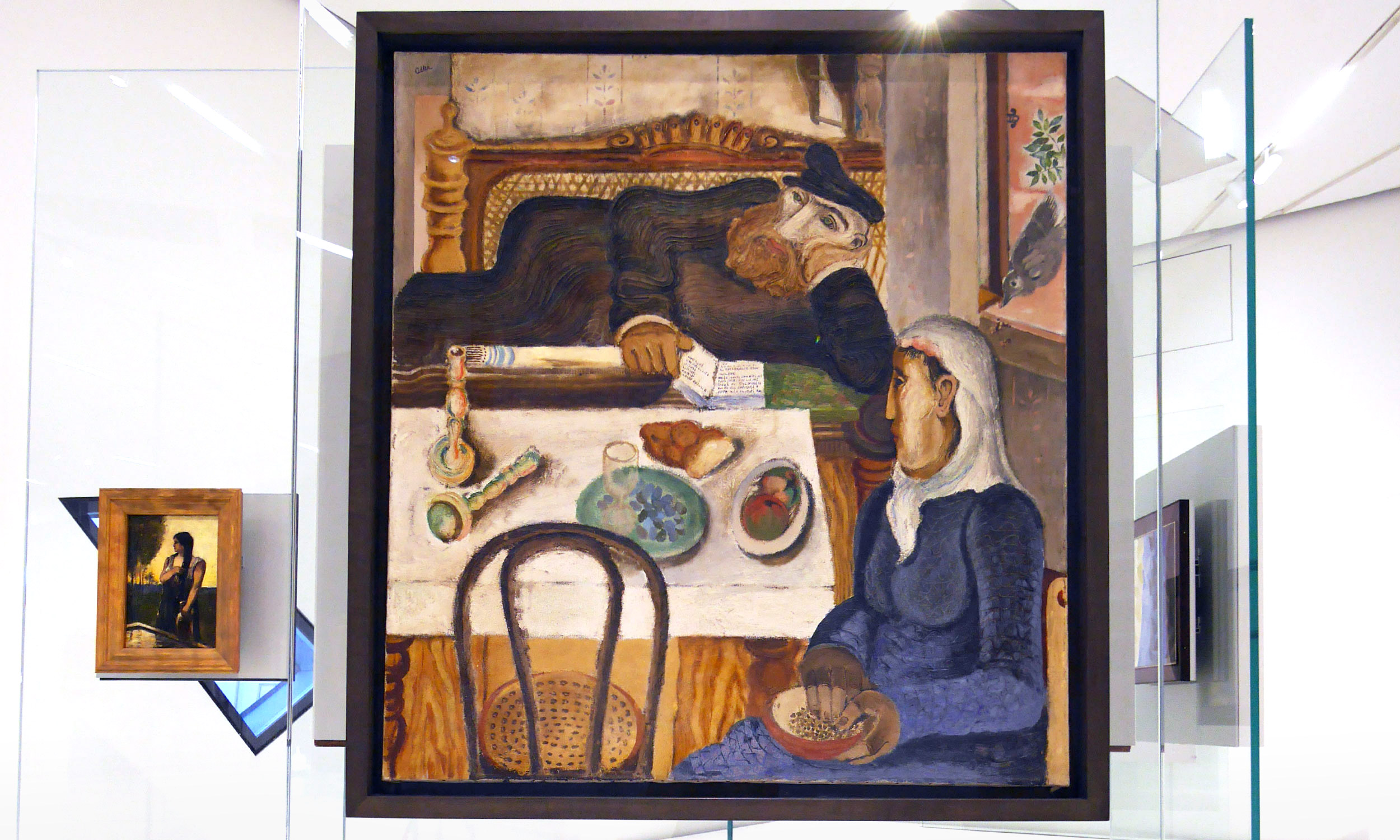 Foto vom Gemälde „Sabbat“ von Janker Adler in Jüdischen Museum, Berlin. Das Kunstwerk hängt an einer Glaswand, die in der Mitte des Raumes steht. Links daneben erkennt man ein weiteres viel kleineres Bild vom selben Künstler, das eine Frau zeigt.