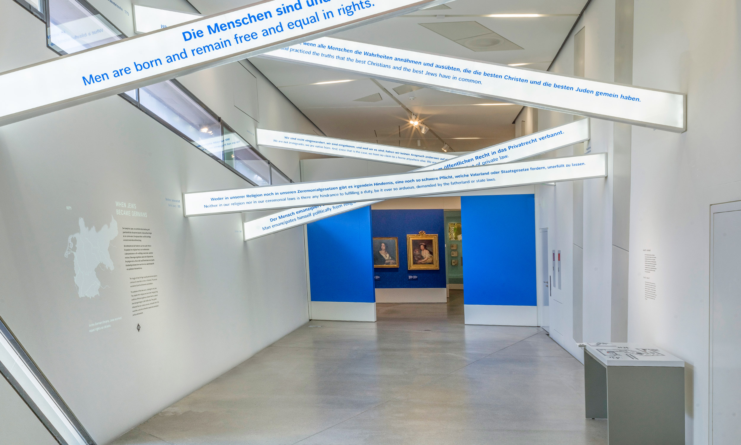 Photo of an interior of the Jewish Museum in Berlin. The room looks like a hallway. At the end of the hallway is an exhibition wall, on which hang two paintings. In the upper part of the room, near the ceiling, there are several white struts with quotes written on them in blue letters on a white background. On the right side, standing against the wall, you can see a tactile plan, which serves the orientation of visually impaired people in the museum building.