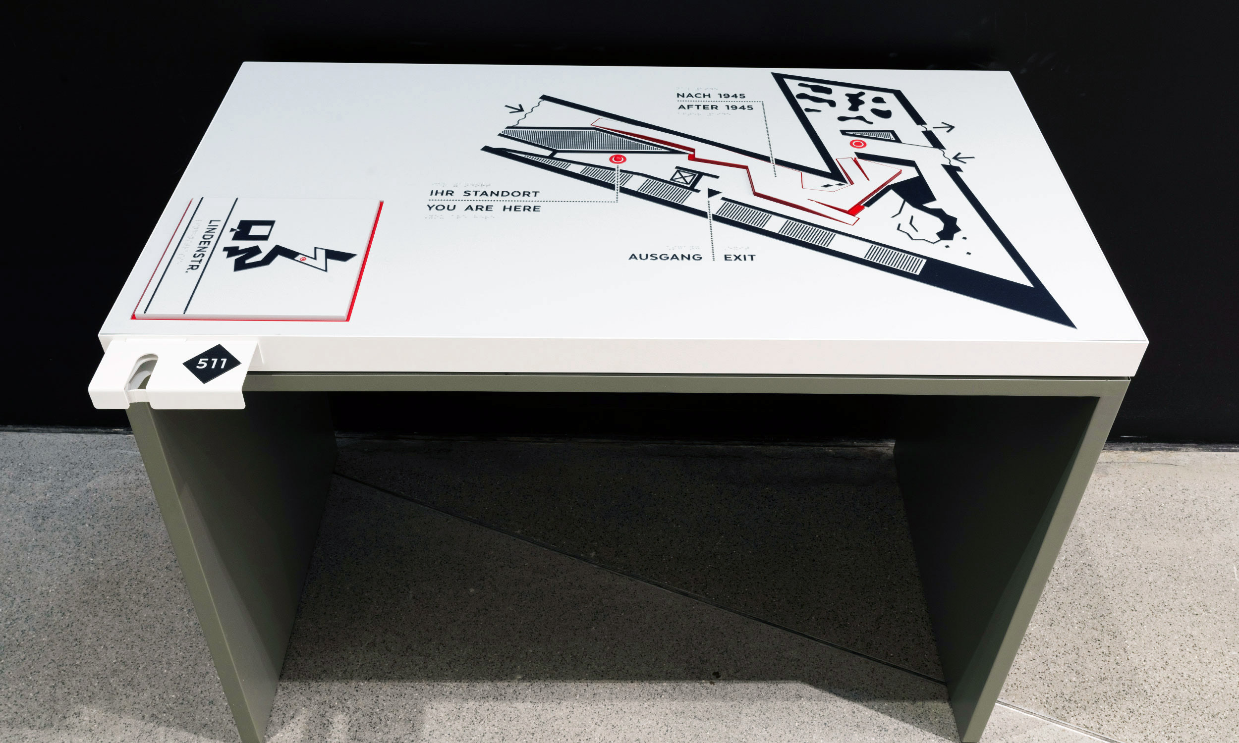 Photo of the tactile map on a table for orientation in the Jewish Museum in Berlin. The tactile map shows on the right side the part of the museum where the visitor is right now. On the left side, smaller, you can see the entire museum building as a tactile plan. On the front left of the table is a cane holder with a number.