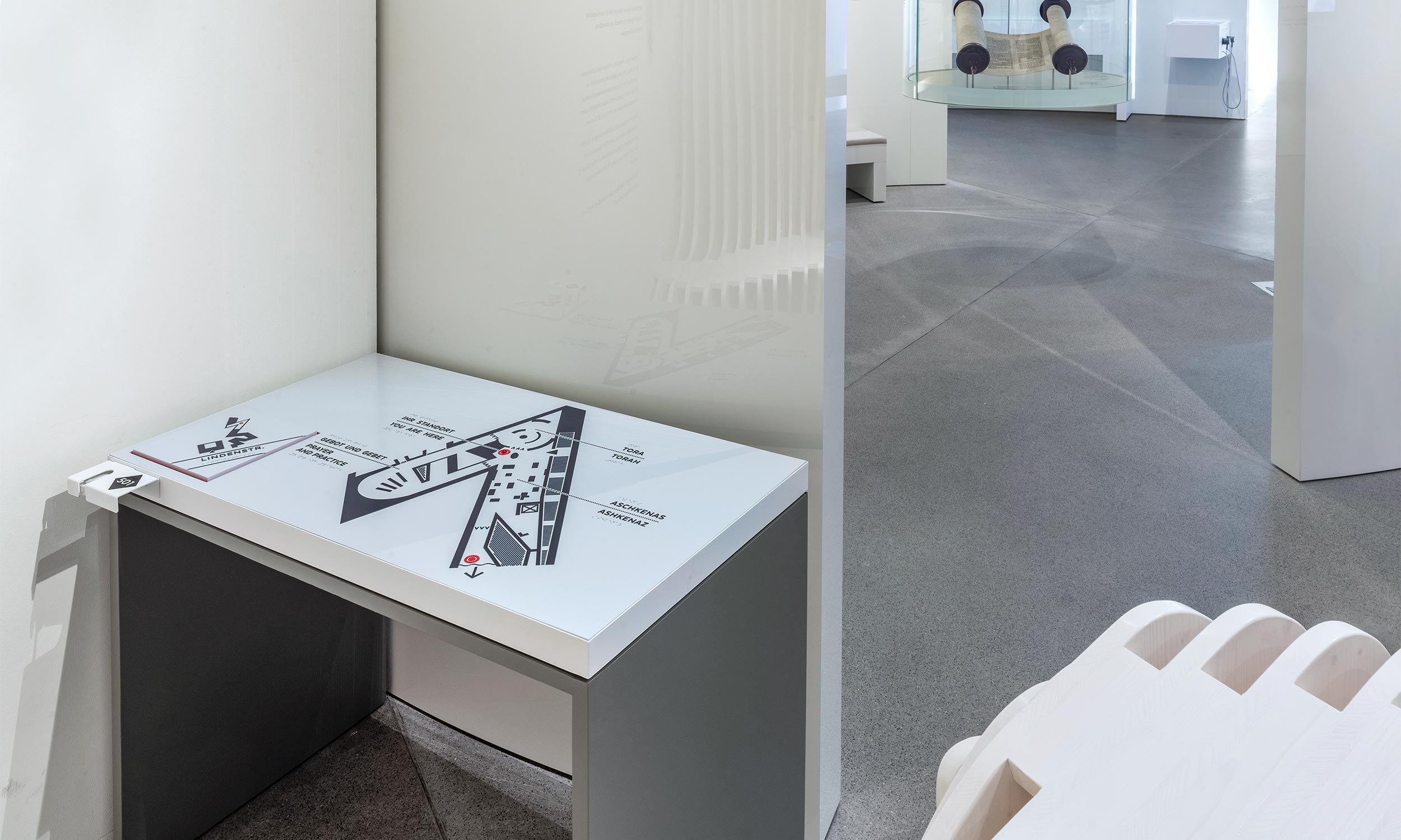 Photo of the tactile map in the Jewish Museum in Berlin fixed on a table. The table stands on the left in a corner which it fills completely. To the right of it, the wall ends and gives a view of the exhibition space with exhibits behind it.