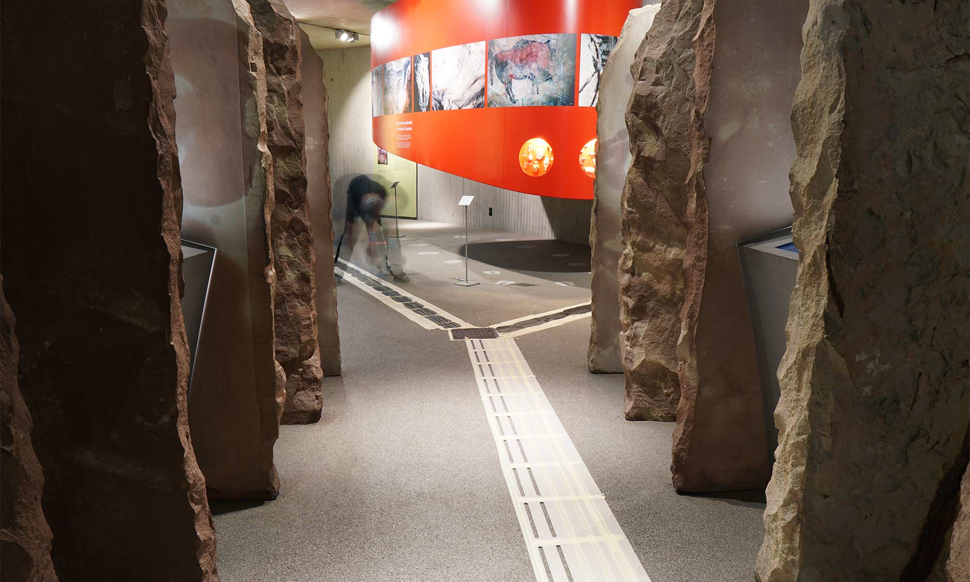 The picture shows the process of laying the floor indicators in the Neanderthal Museum. On the sides of the picture, you can see partitions that have been set up to look like rocks. A path runs straight through the picture and branches to the left and right at one point. The indicators are still taped off. In the back of the picture, you can see an installer bending down and pouring black resin onto the masked surfaces.