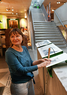 Tamara Ströter reads Braille on a tactile model. This tactile model is a shape bent from sheet steel, which is mounted on a wall offset at a slight angle. In the background you can see parts of the exhibition and a staircase leading up several floors of the museum.