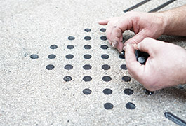 Ein Monteur klebt per Hand kleine, schwarze Punkte für ein quadratisches Aufmerksamkeitsfeld der Bodenindikatoren.