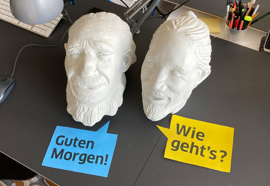 Two white models of Neanderthal heads placed on a desk in the inkl.design office. In front of them is colorful paper cut out into speech bubbles with the words: Good morning! and How are you?
