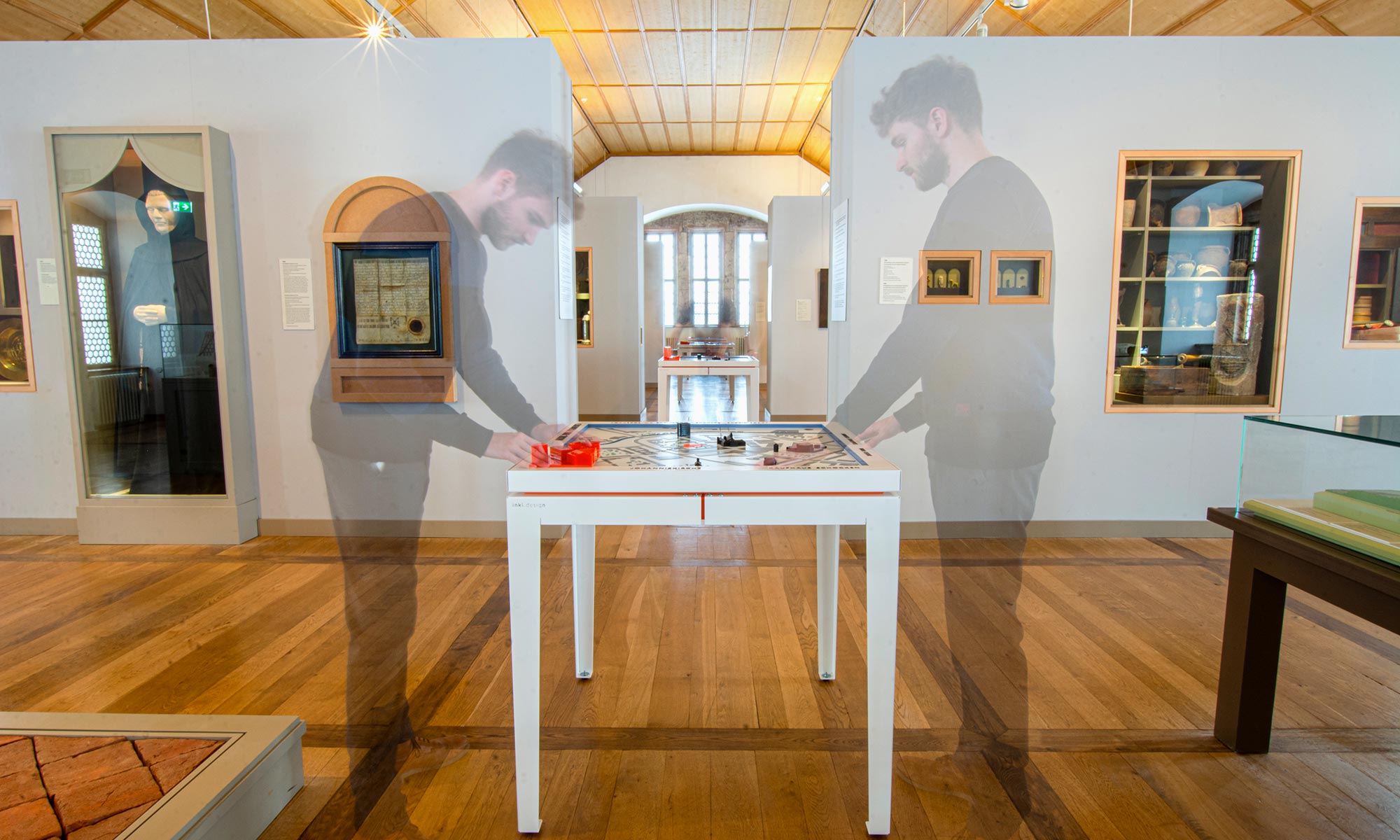 The photo shows an exhibition room of the Schlossbergmuseum Chemnitz. The floor and ceiling are made of wood, white walls divide the large room into different areas. In the foreground, one of the four tactile models stands on a white metal table. A visitor can be seen shadowily to the left and right of the table. He is touching the tactile model of the city.