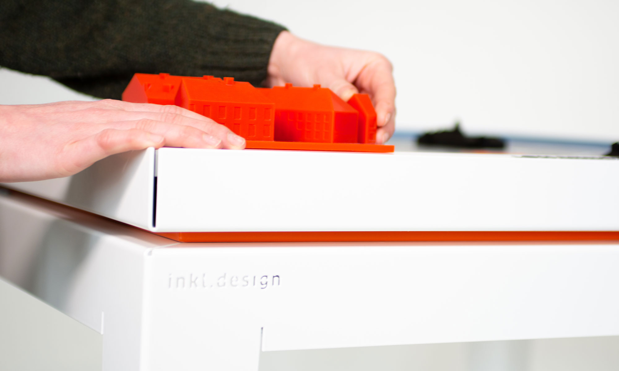 A close-up shows two hands touching the corner of the model where a three-dimensional detail of the building can be found. The base is clearly recognisable: a white metal table with the engraving "inkl. design", on top of which is a likewise white metal tray that supports the model. Between the two is an orange board.