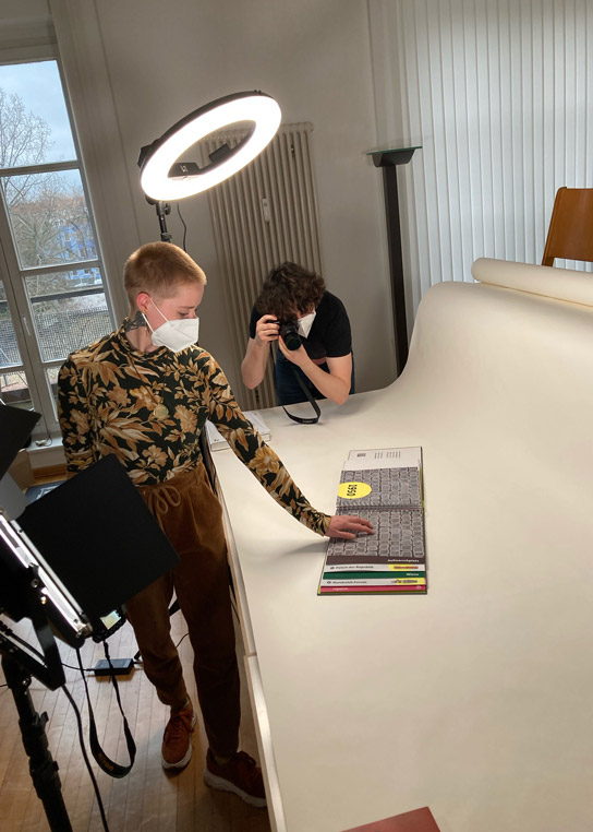 Makin-of Aufnahme aus dem inkl-Design Büro. Zwei Mitarbeiter*innen fotografieren das Tastbuch. Im Raum ist eine professionelle Beleuchtung mit Scheinwerfern und Ringlicht aufgebaut.