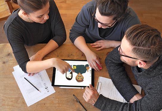 Drei Mitarbeiter von inkl.Design sitzen an einem Tisch und gucken auf eine Illustration auf einem Tablet.