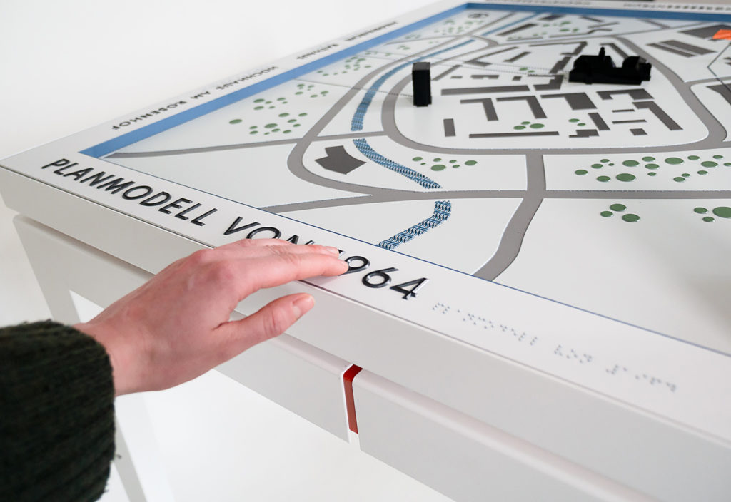 Das Projektbild zeigt eine Hand, von links ins Bild kommend, betastet die Beschriftung des taktilen Modells der Planstadt von 1964.