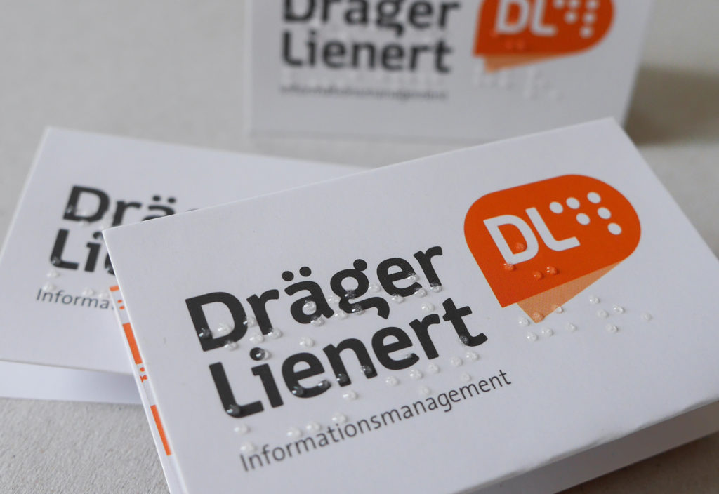 Photo with inclusive business cards from Draeger & Lienert. The business cards are folded in three parts. The standard-compliant and easily tactile Braille is applied to the pages with transparent varnish.