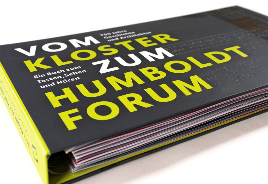 Studio shot of the closed tactile book "From the Monastery to the Humboldt Forum" on a white background. The focus of the image is on the title of the book in bold capital letters. These were depicted in bright neon yellow and are printed on the dark gray background of the cover.