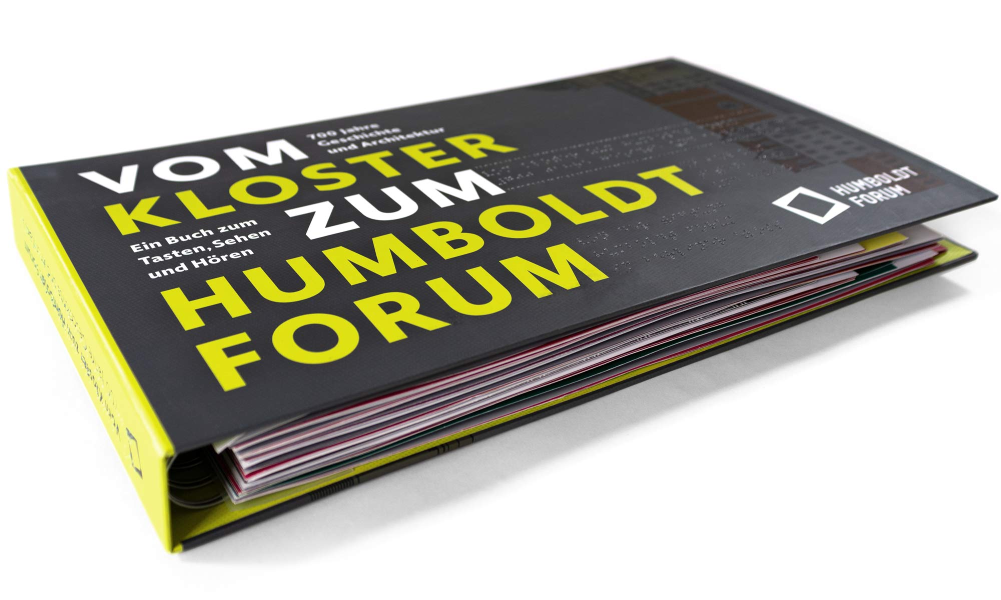 Studio shot of the closed tactile book "Vom Kloster zum Humboldt Forum" on a white background. The focus of the image is on the title of the book written in bold capitals. These were depicted in bright neon yellow and are printed on the cover's dark grey background.