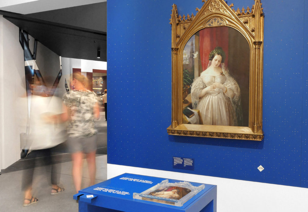 The project picture shows Till Henning and another employee of inkl.Design together with Tamara Ströter at a tactile model in the Neanderthal Museum. In the center is the painting -Albertine Heine als Braut. The painting hangs on a blue wall. The oil painting shows Albertine Albert in a wedding dress in front of a red, slightly opened curtain. In front of the artwork is the accompanying tactile model including explanation in Braille. Two visitors can be seen out of focus in the background on the left.